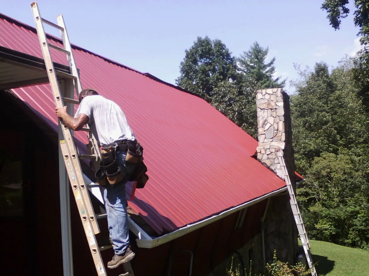 Roofer on a ladder beside a red metal roof and stone chimney — Soffit & Fascia Repair by Roofing Brothers in Dunnellon